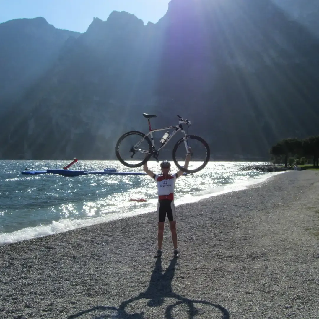 Christian Zoss hebt sein Rad hoch vor einer Bergkulisse am See